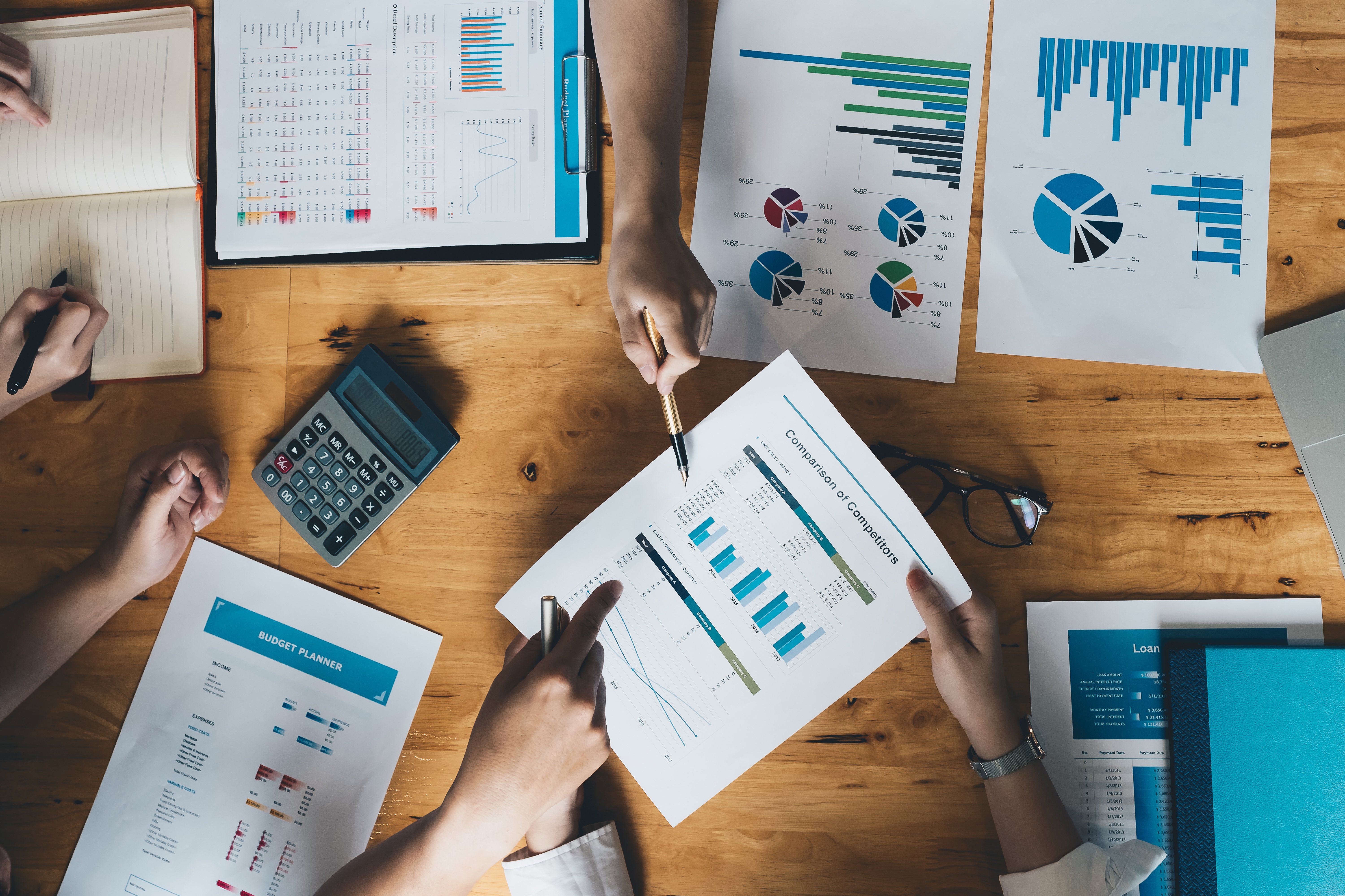 Differentiation Beyond PPC: Why Brand Strategy Multiplies Ad Performance 1 A group of people collaborating on financial and marketing analysis at a wooden table covered with charts, graphs, and reports showing data comparisons, with calculators, notebooks, and pens in use during the discussion.