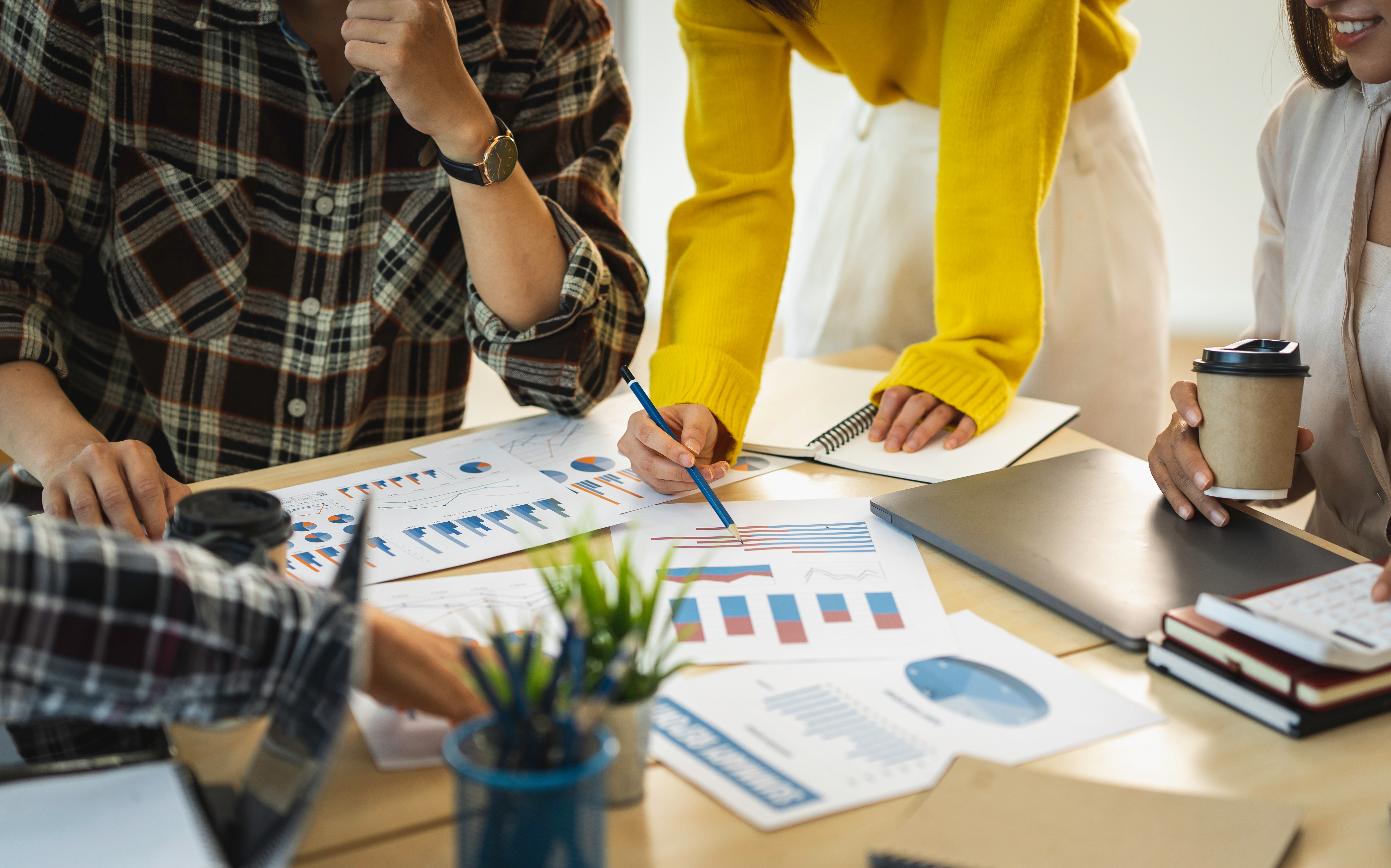 Branding as the Law Firm Growth Strategy of 2026 1 Close-up of a diverse team collaborating at a wooden table, reviewing printed charts and graphs with colorful bar and pie diagrams. One person in a plaid shirt is thinking while another in a bright yellow sweater points to a document with a pencil. Other team members hold a coffee cup and a calculator, surrounded by a laptop, notebooks, and a small potted plant.